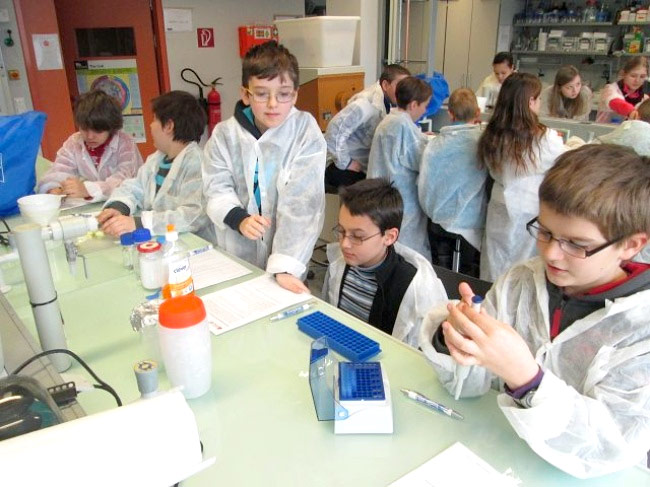 Viele Kinder um einen Tisch mit weißen Laboranzügen